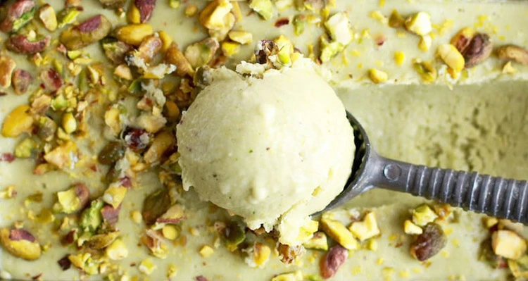 The photo shows a scoop of pistachio frozen yogurt drawn from a pistachio ice cream tray. The ice cream is drizzled with pistachio pieces.